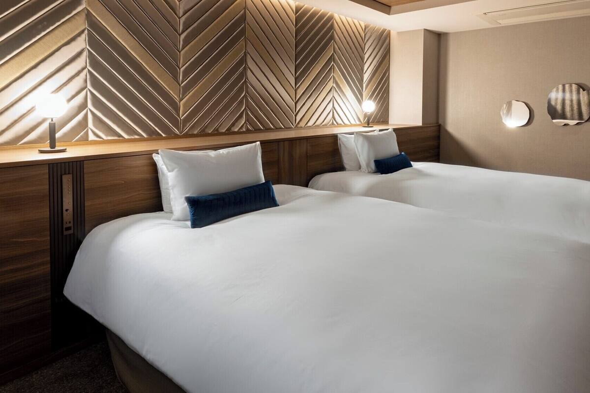 Quarto de hotel com duas camas, uma cabeceira de madeira com padrão em zigue-zague e luminárias fixadas na parede.
