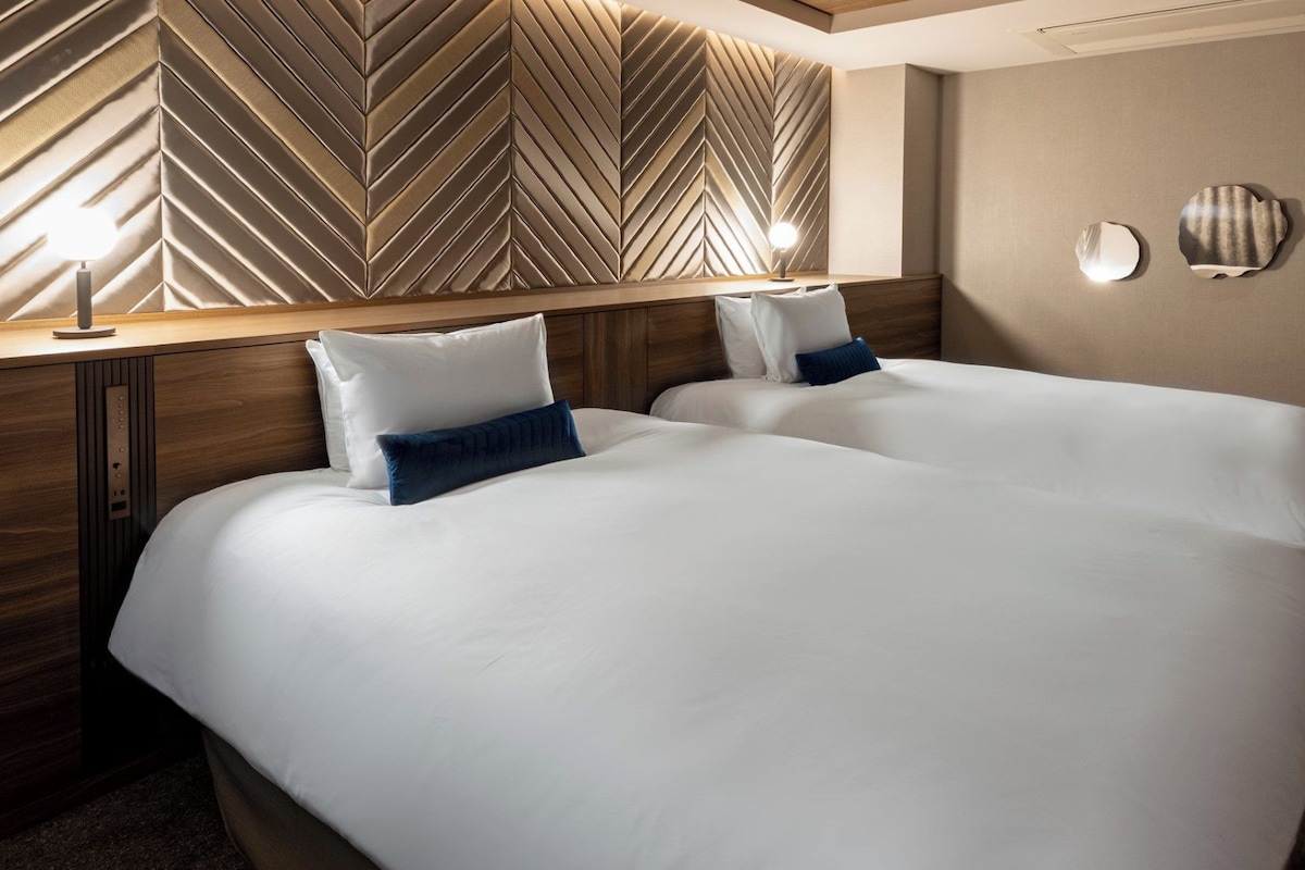 A hotel room with two beds, a wooden headboard with a chevron pattern, and wall-mounted lights.