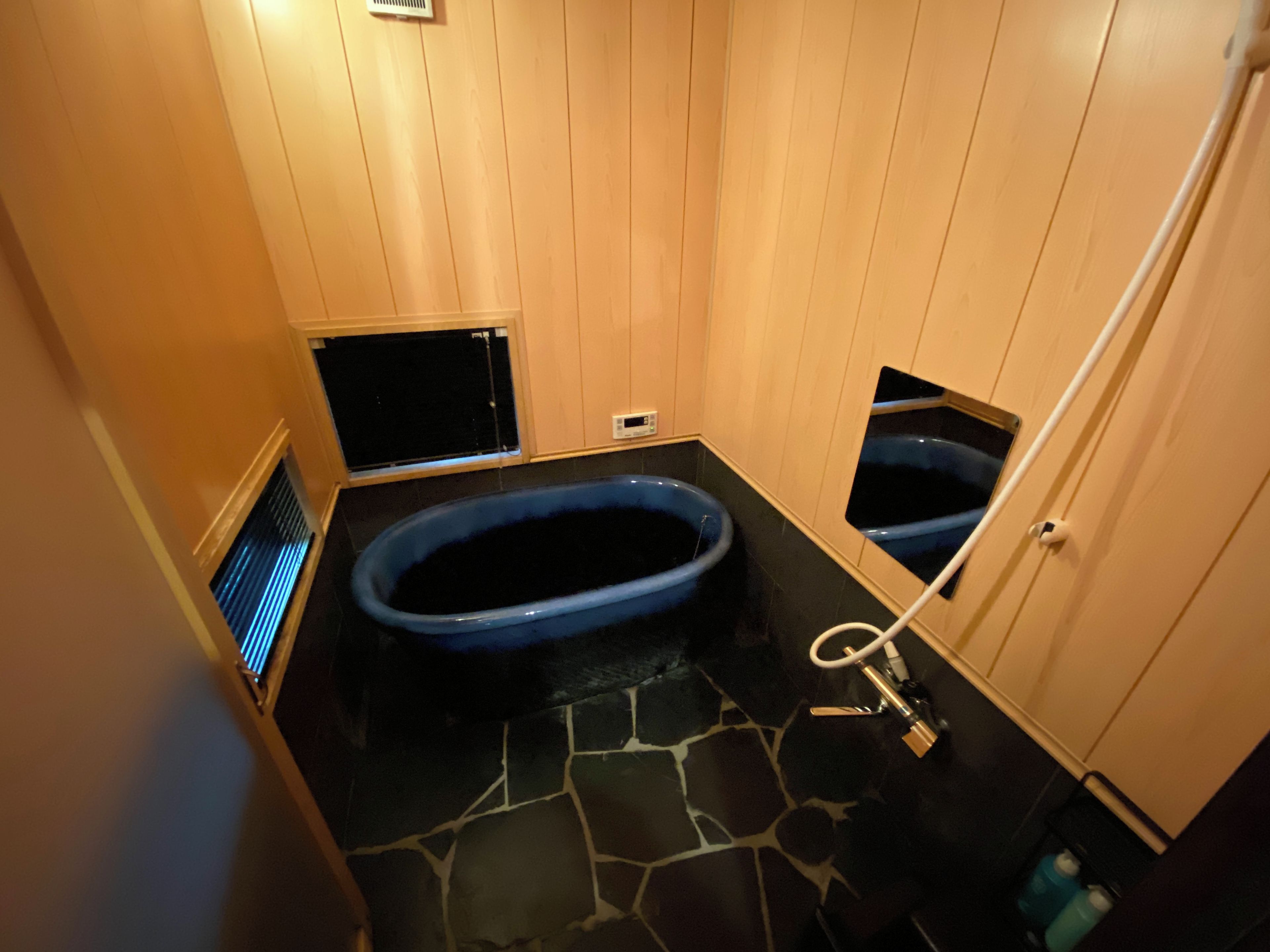 A small, dimly lit bathroom with a blue bathtub, a stone floor, and wooden walls.