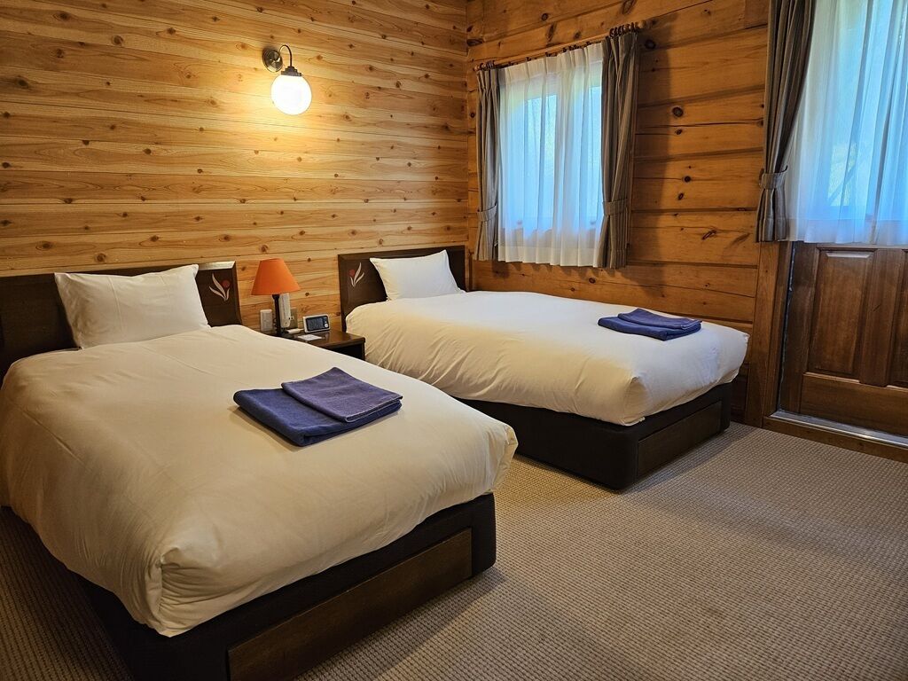 Una habitación con dos camas, paredes de madera, una mesita de noche con lámpara y una ventana con cortinas.