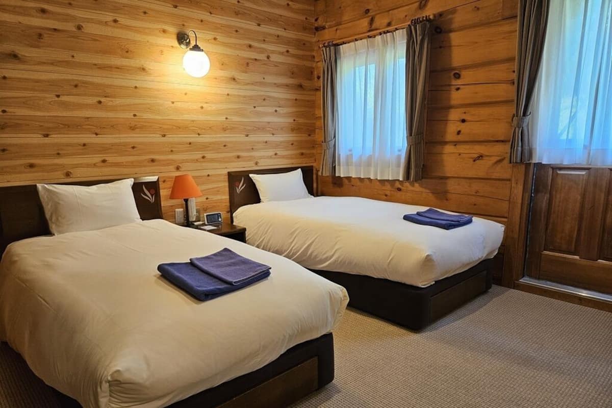 Habitació amb dos llits, parets de fusta, una cambrera amb una làmpada i una finestra amb cortines.