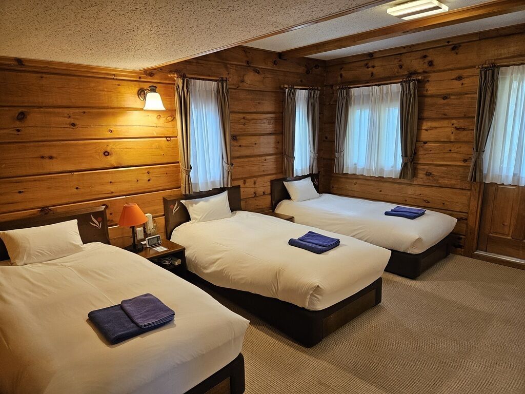 Una habitación con tres camas, paredes de madera y una mesita de noche con lámpara.