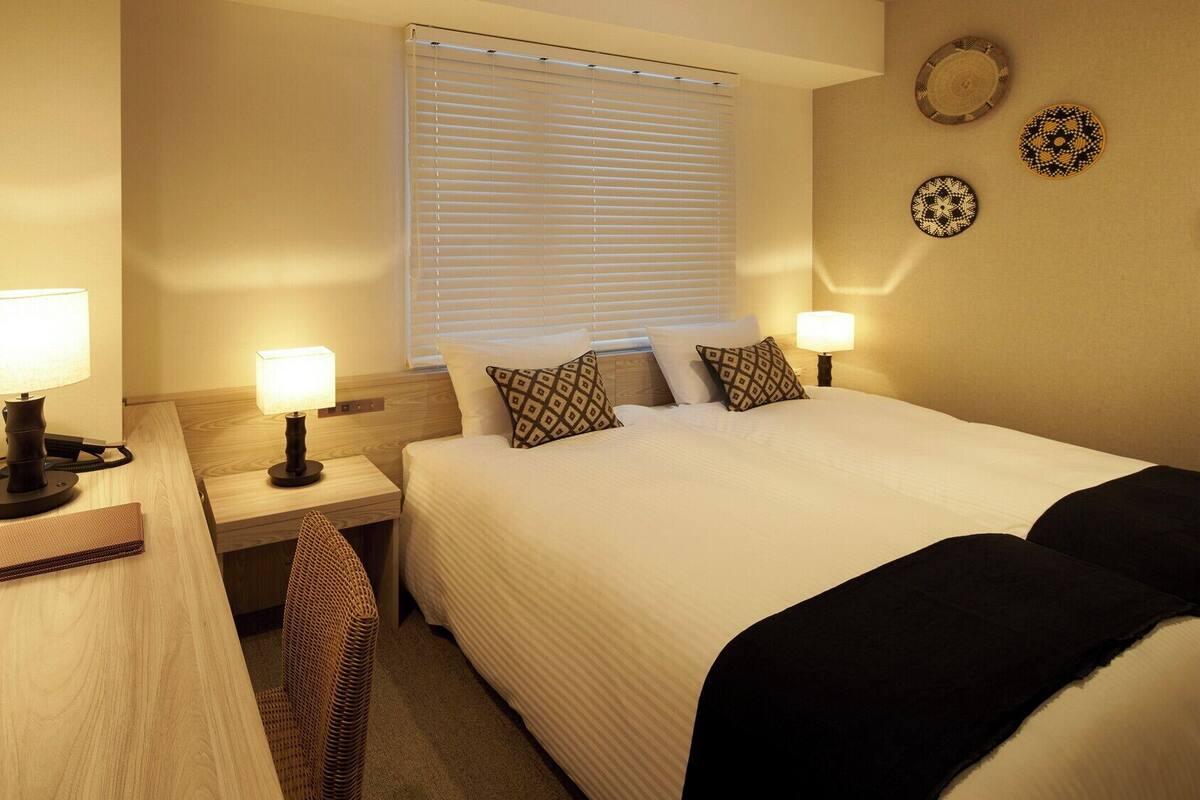 A hotel room with a bed, bedside lamps, a mirror, a window with blinds, and decorative plates on the wall.