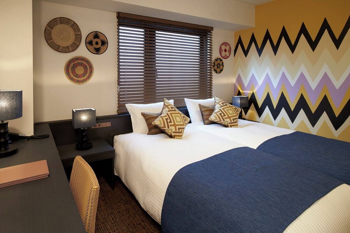 A hotel room with a bed, two bedside tables, a desk, a chair, and a window with blinds.