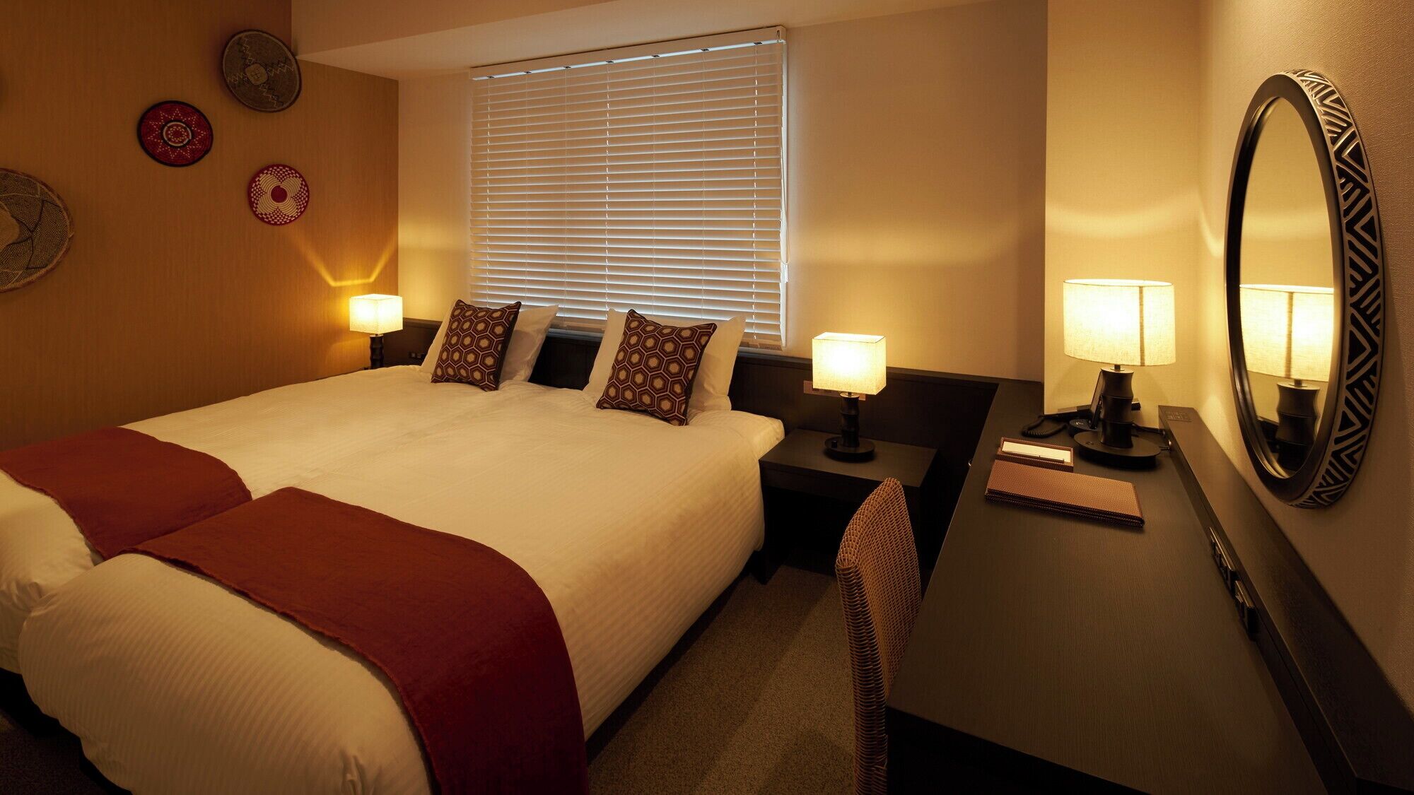 A hotel room with a large bed, two bedside tables with lamps, a desk with a mirror, and a window with blinds.