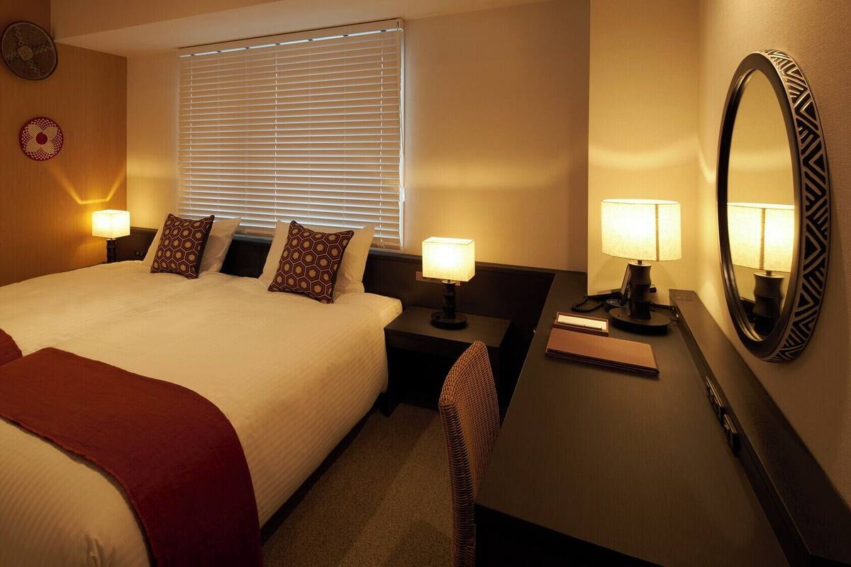 A hotel room with a large bed, two bedside tables with lamps, a desk with a mirror, and a window with blinds.