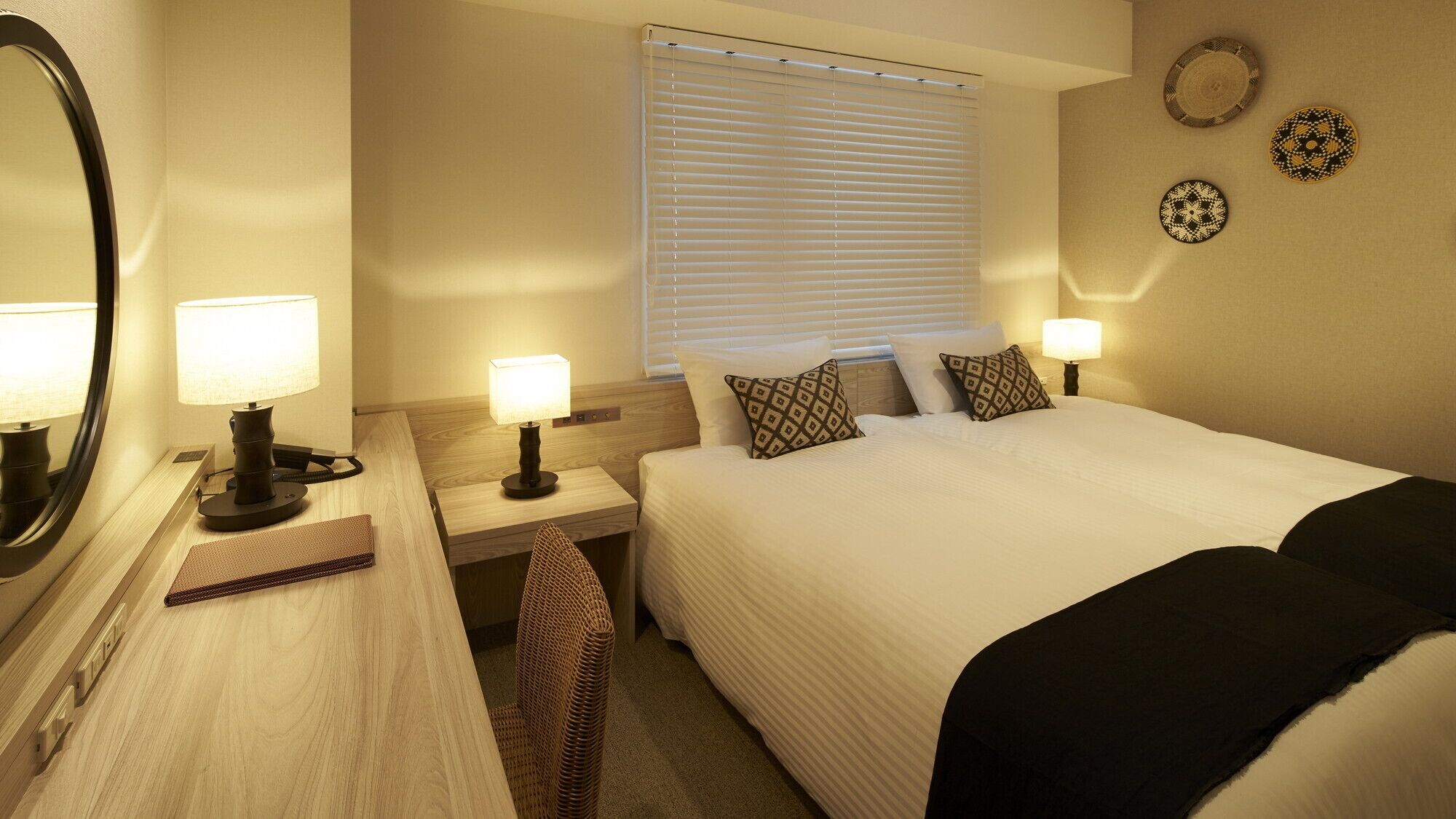 A hotel room with a bed, bedside lamps, a mirror, a window with blinds, and decorative plates on the wall.