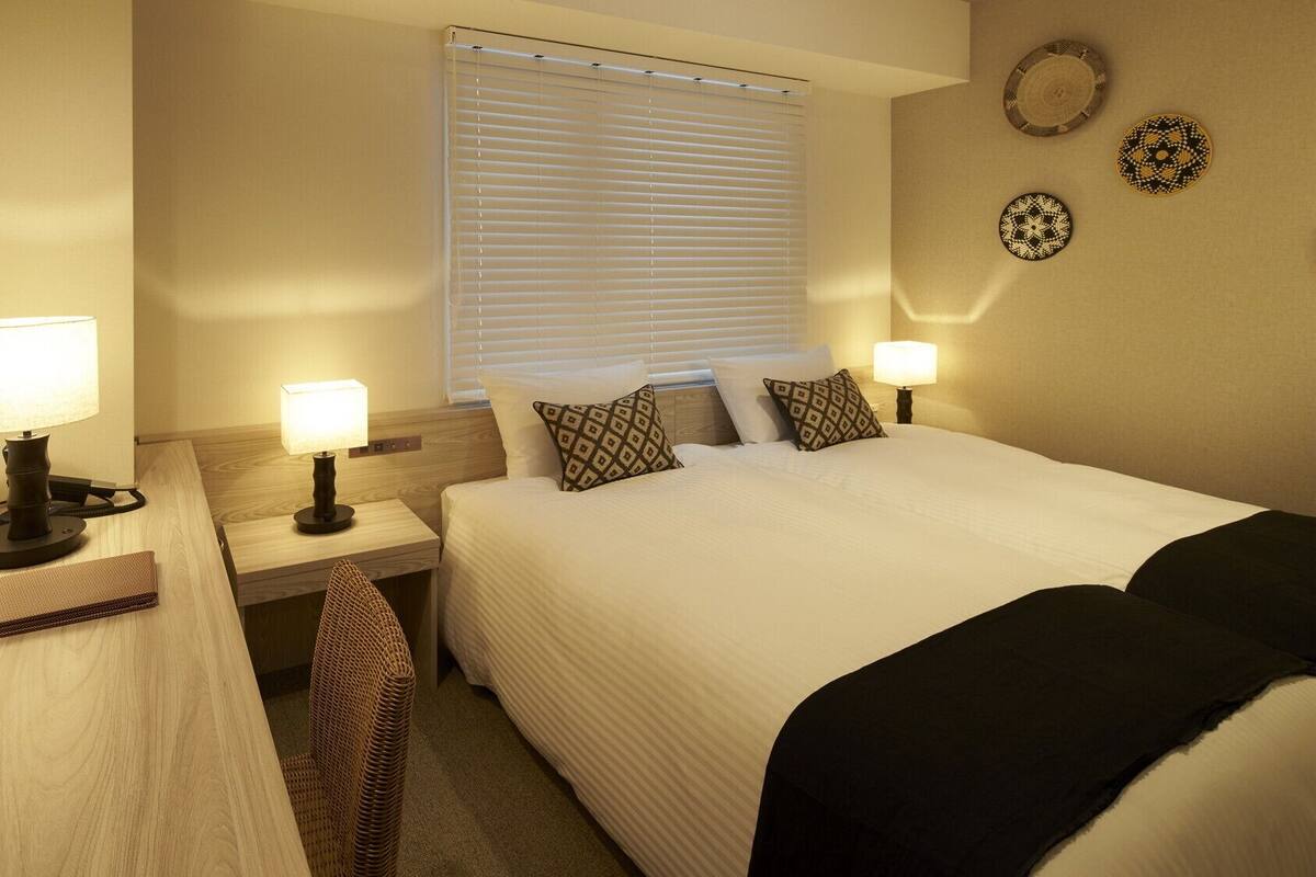 A hotel room with a bed, bedside lamps, a mirror, a window with blinds, and decorative plates on the wall.