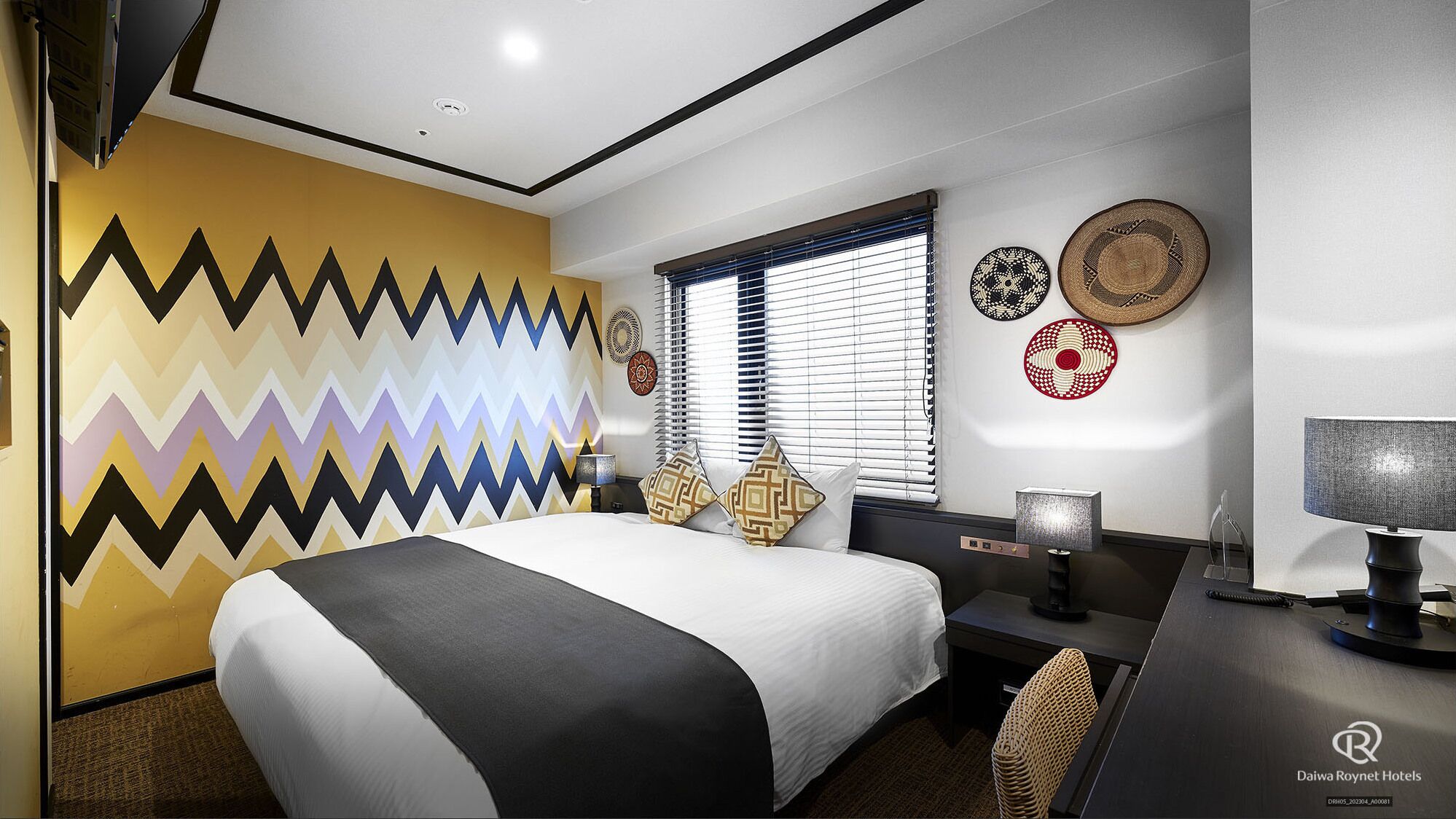 A hotel room with a bed, desk, and window with blinds.