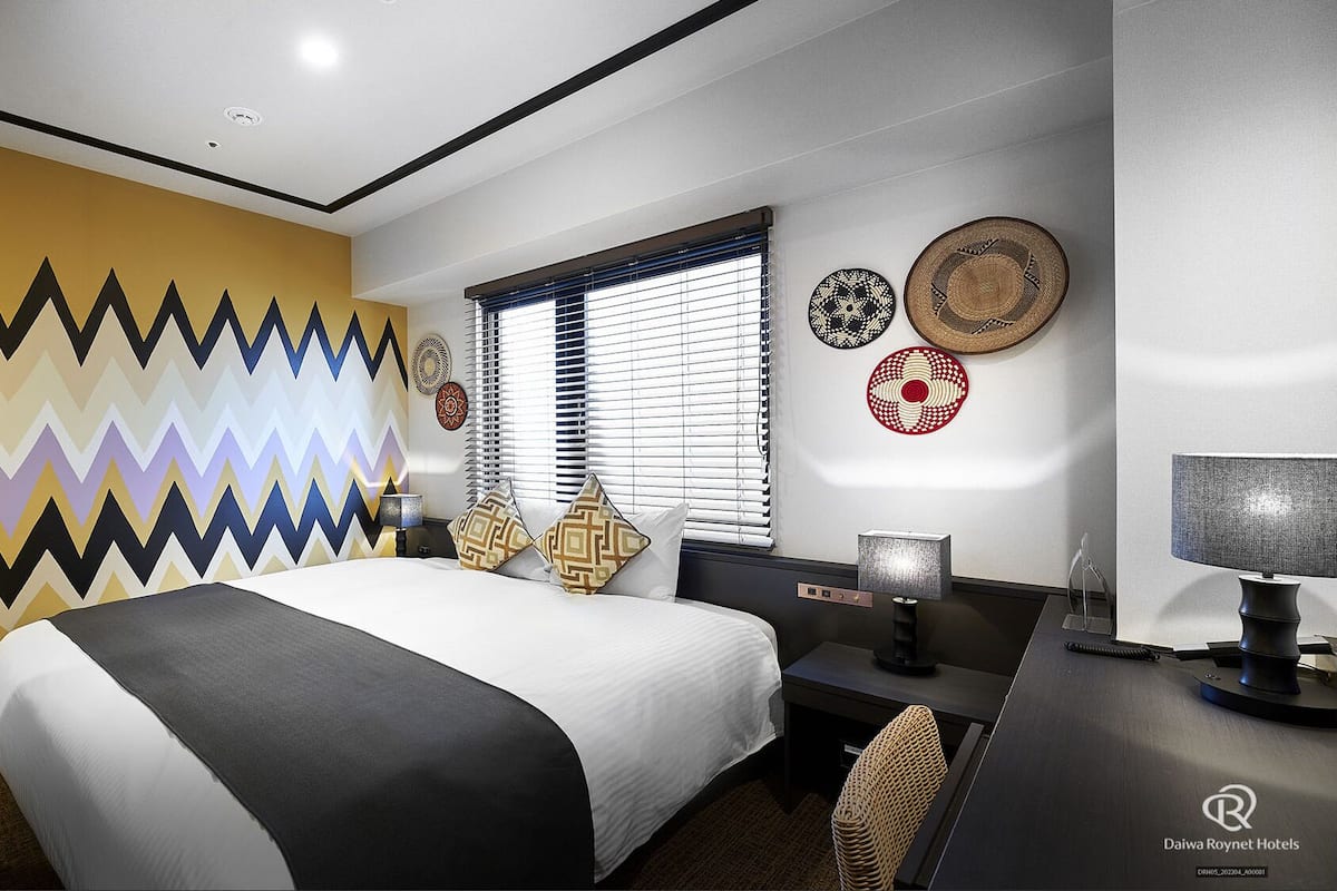 A hotel room with a bed, desk, and window with blinds.