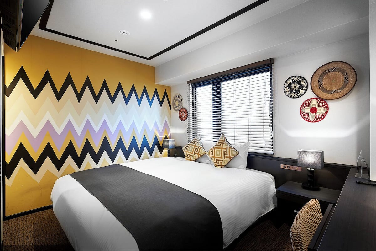 A modern hotel room with a large bed, a desk, and a window with blinds.