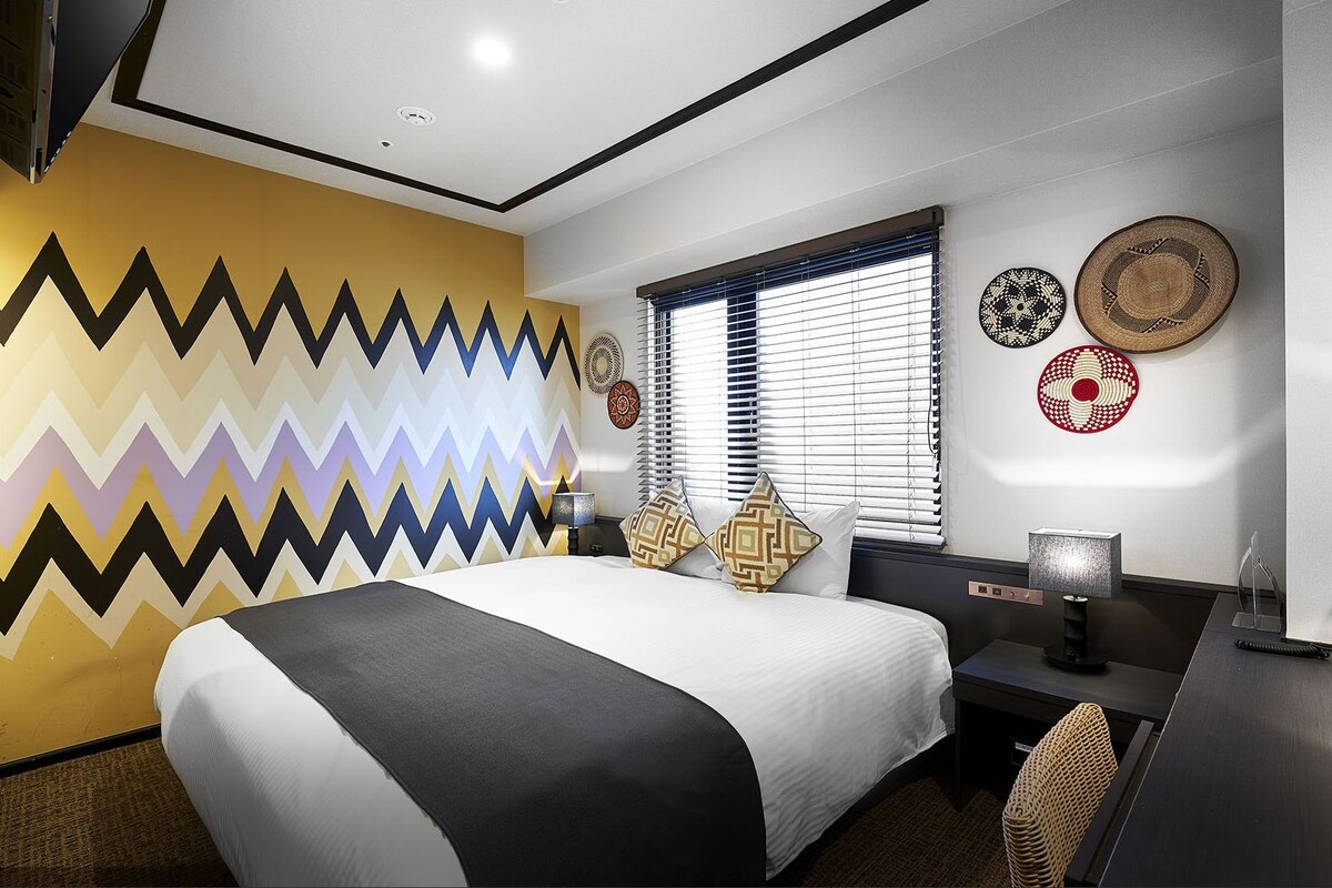 A hotel room with a bed, desk, and window with blinds.