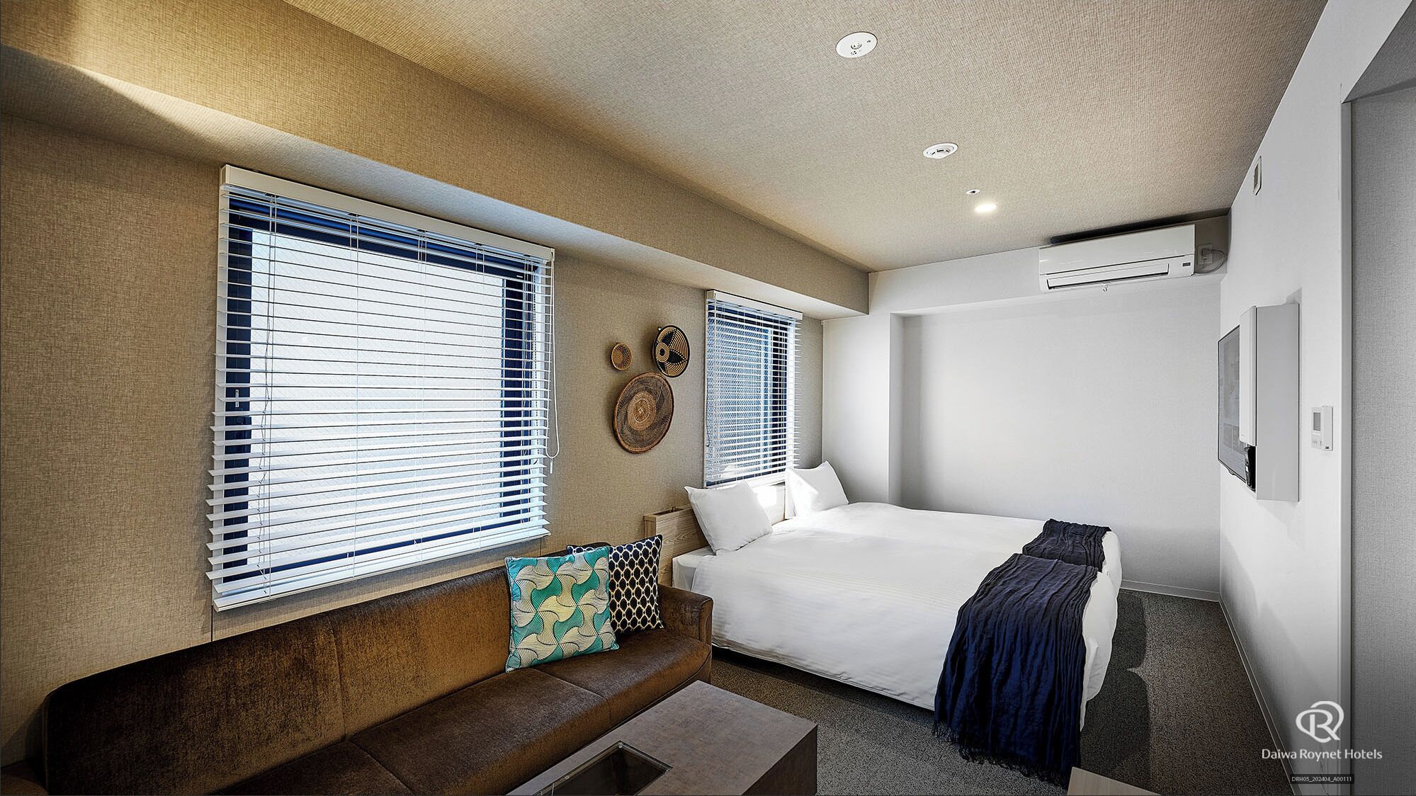 A modern hotel room with a bed, a sofa, and two windows with blinds.