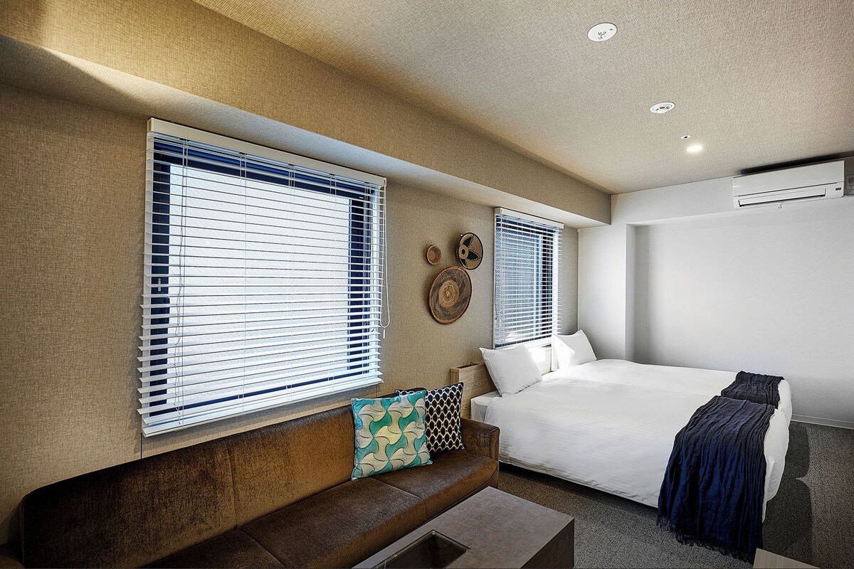 A modern hotel room with a bed, a sofa, and two windows with blinds.