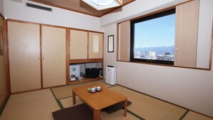 Ein kompakter Raum mit Tatami-Boden, einem Holztisch, einem Fernseher und einem Fenster mit Blick auf die Stadt.
