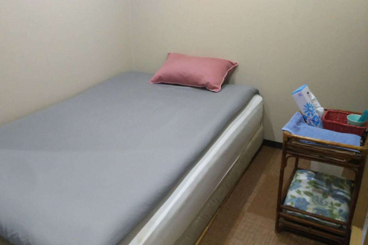 A single bed with a grey mattress and a pink pillow, a small wooden nightstand with a basket and a magazine, and a plain wall.