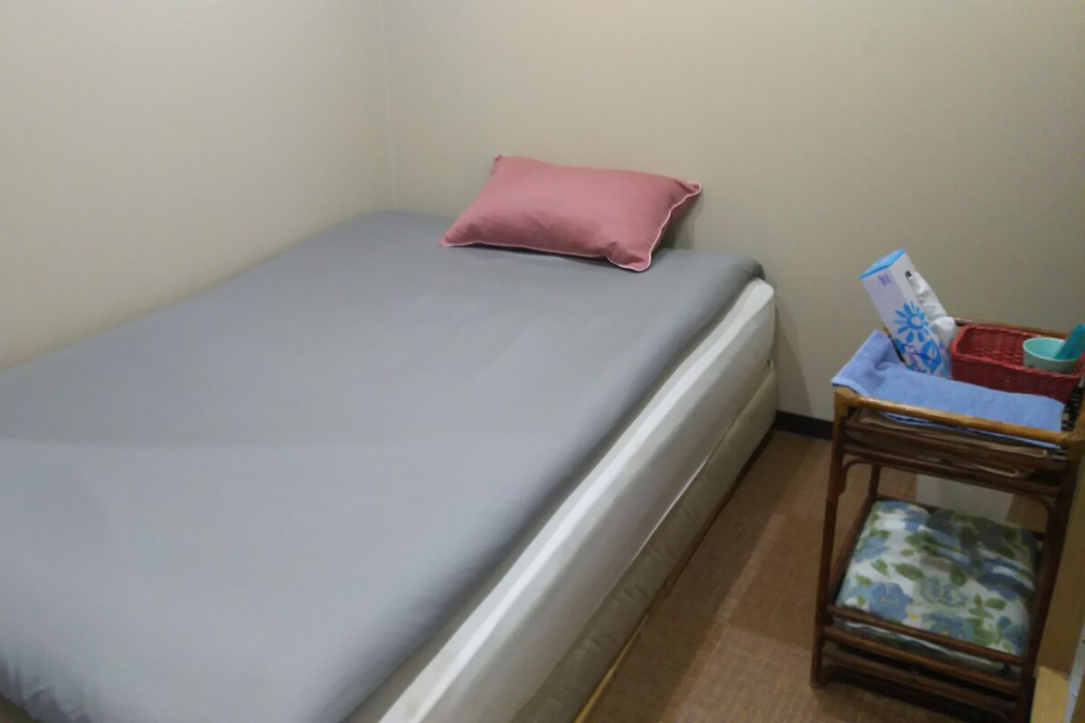 A single bed with a grey mattress and a pink pillow, a small wooden side table with a basket, and a chair with a floral cushion.