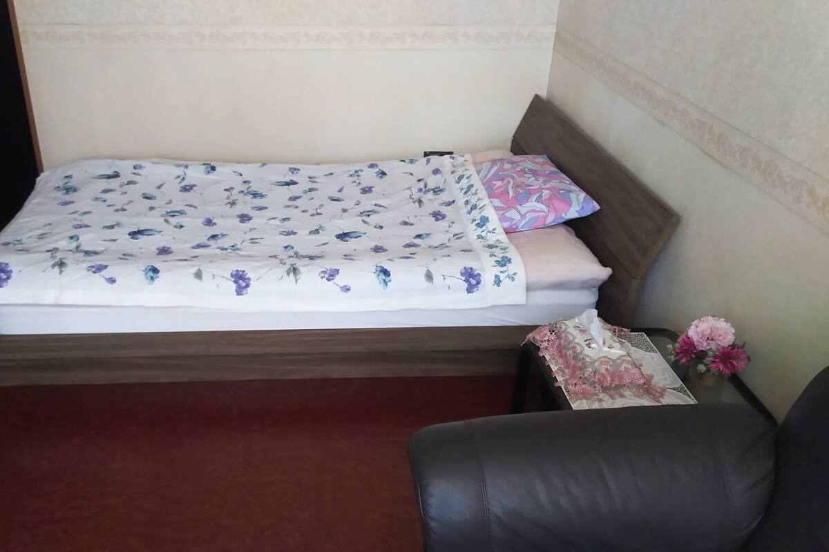 A bed with a floral bedspread, a pillow, a bedside table with a vase of flowers, and a leather chair.