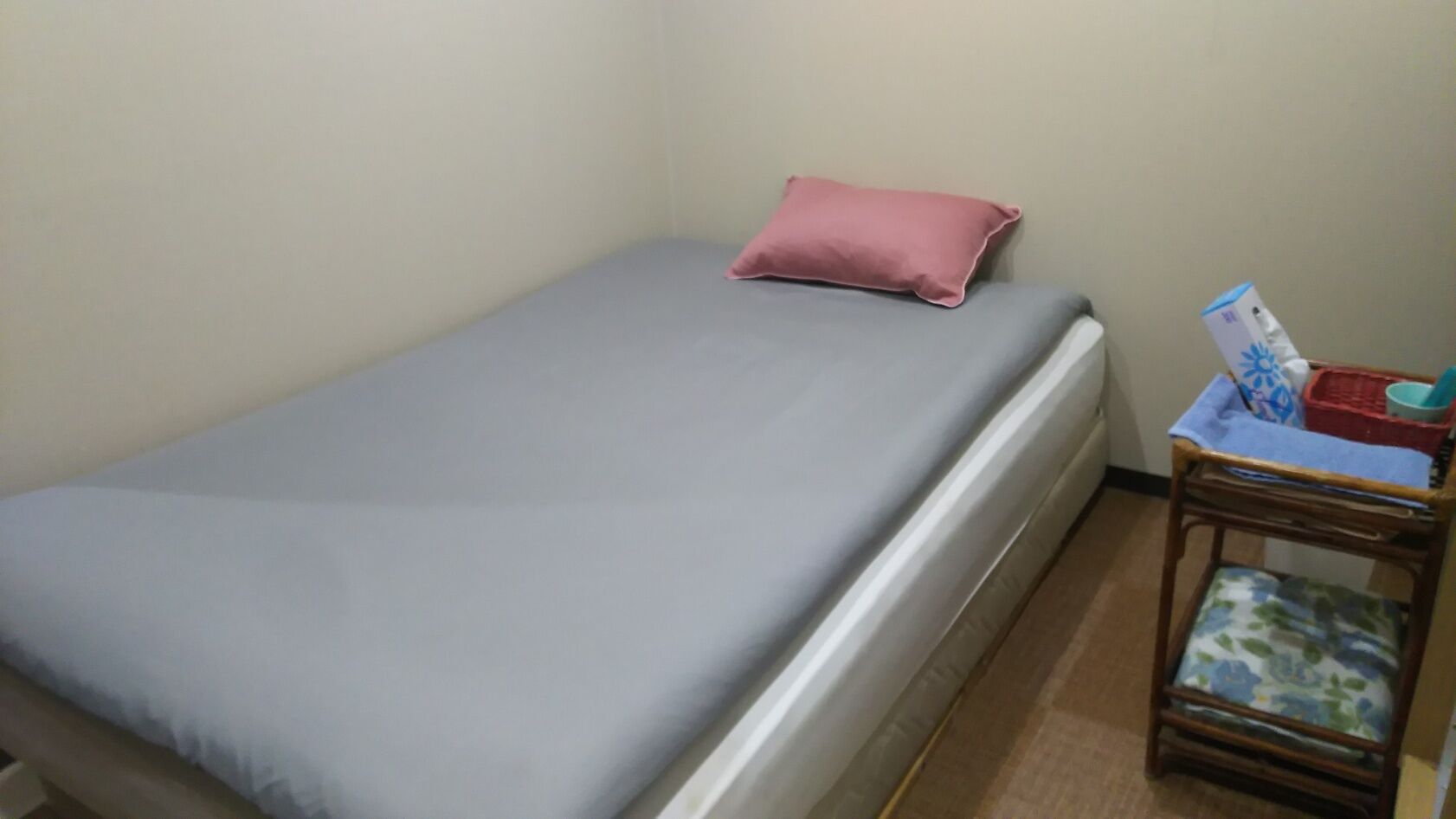 A single bed with a grey mattress and a pink pillow, a small wooden nightstand with a basket and a magazine, and a plain wall.