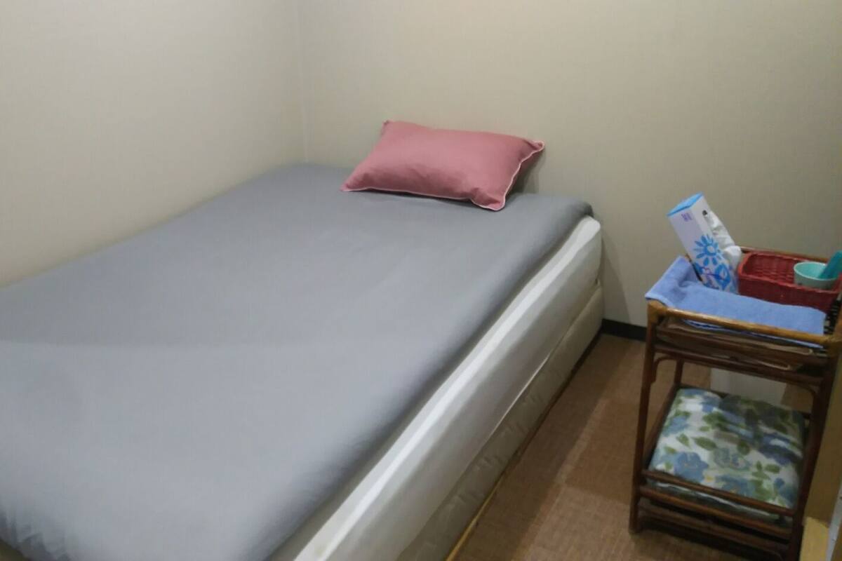 A single bed with a grey mattress and a pink pillow, a small wooden nightstand with a basket and a magazine, and a plain wall.