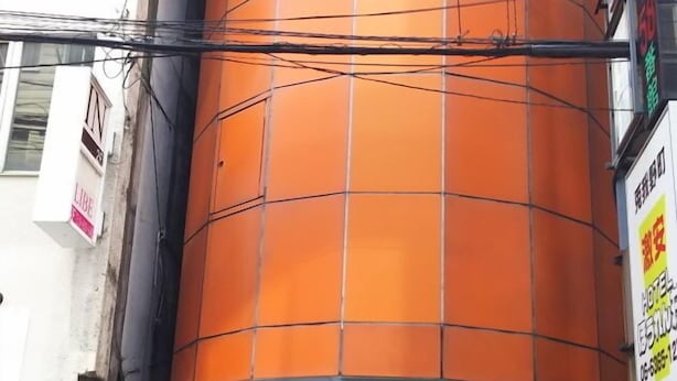 A building with a large orange cylindrical structure, a sign reading 'OSAKAYA 58 BAN-KAN,' and various signs and advertisements around the entrance. - Hotel 4F Macaroni (Osaka)