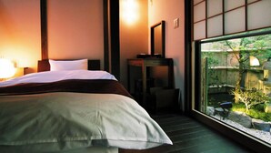 Een slaapkamer met een bed, nachtkastje, lamp en uitzicht op een tuin door een groot raam.