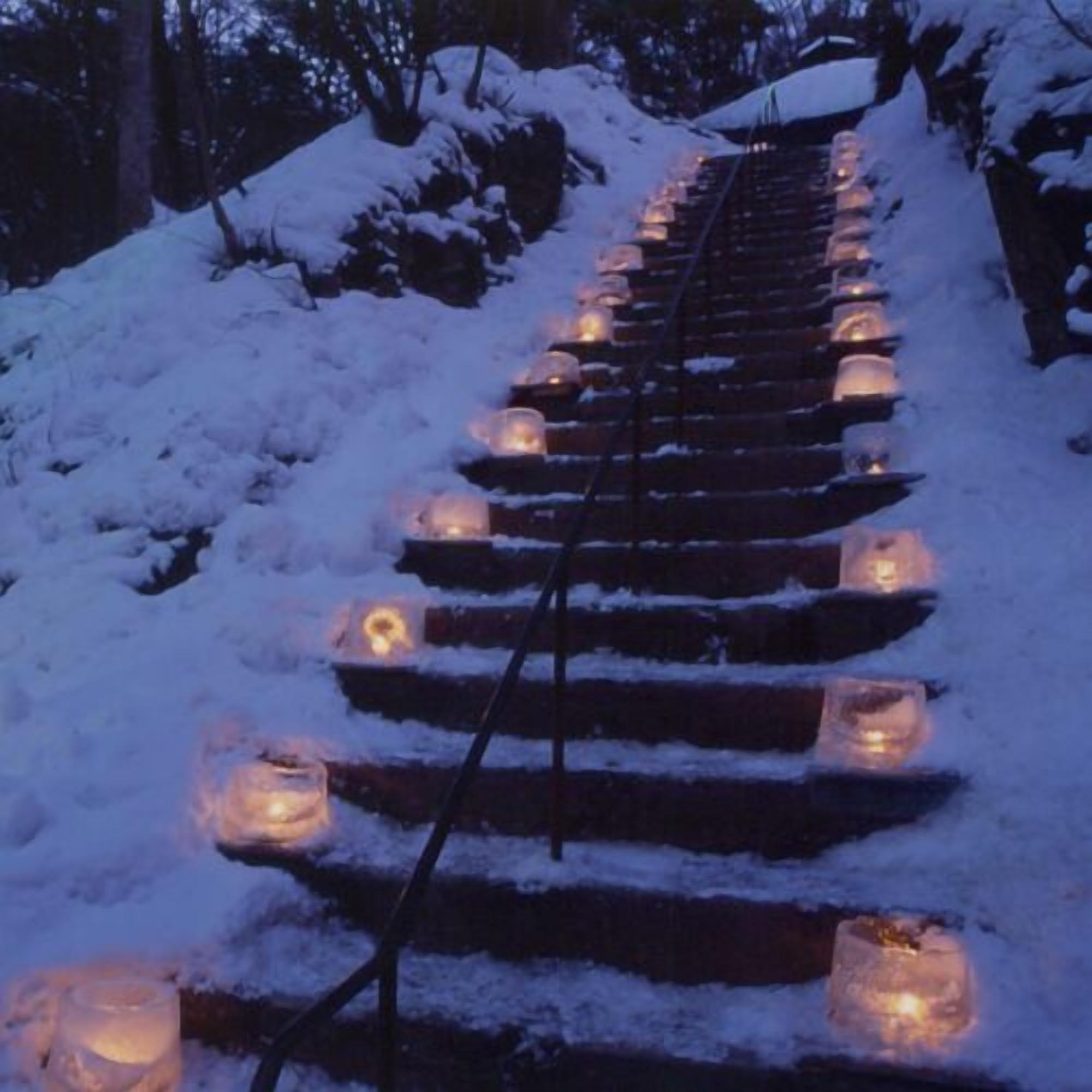 Eine mit Schnee bedeckte Treppe, an deren Stufen in Abständen Kerzen stehen.