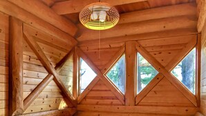 Interieur van een blokhut met een schuine balken plafond, houten wanden en een raam met een kruisverbinding.