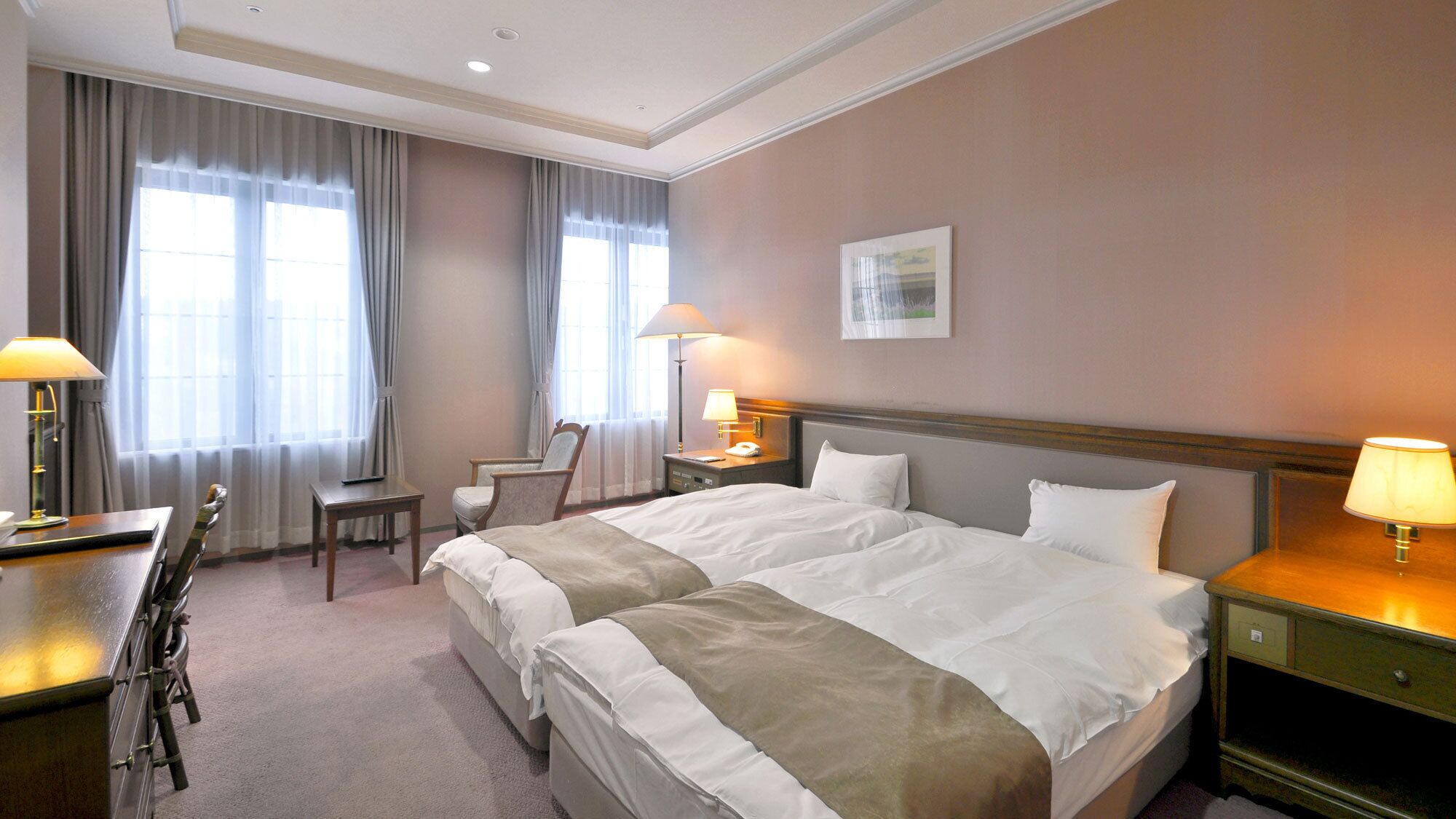 A hotel room with a double bed, a desk with a chair, a bedside table with a lamp, and a window with curtains.