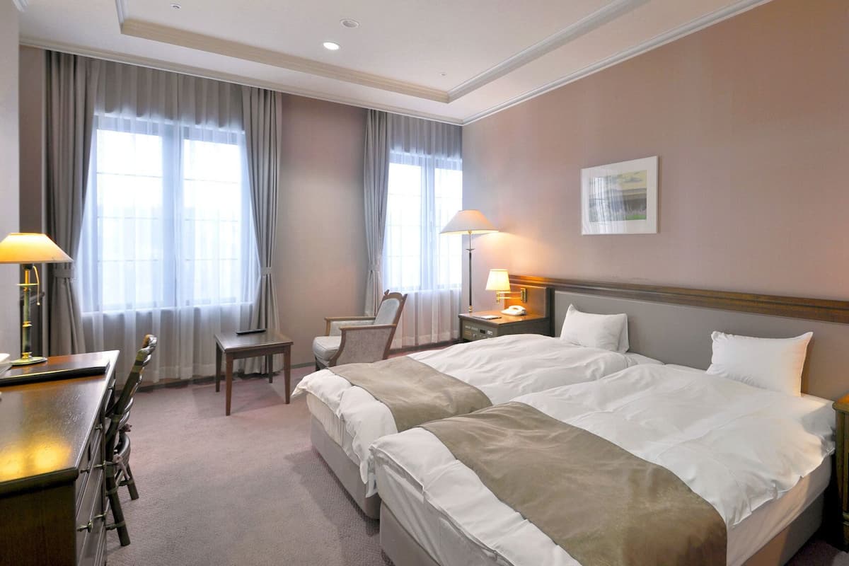 A hotel room with a double bed, a desk with a chair, a bedside table with a lamp, and a window with curtains.