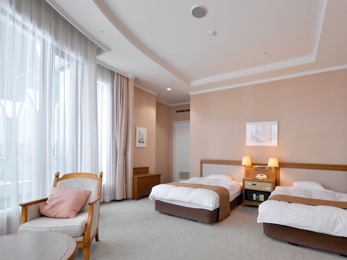 A hotel room with two beds, a chair, a table, and a window with curtains.