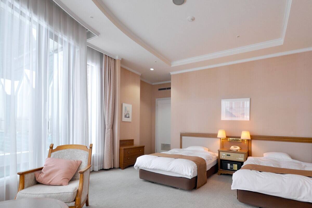 Hotellihuone, jossa on kaksi sänkyä, tuoli, pöytä ja ikkuna, jossa on verhot.