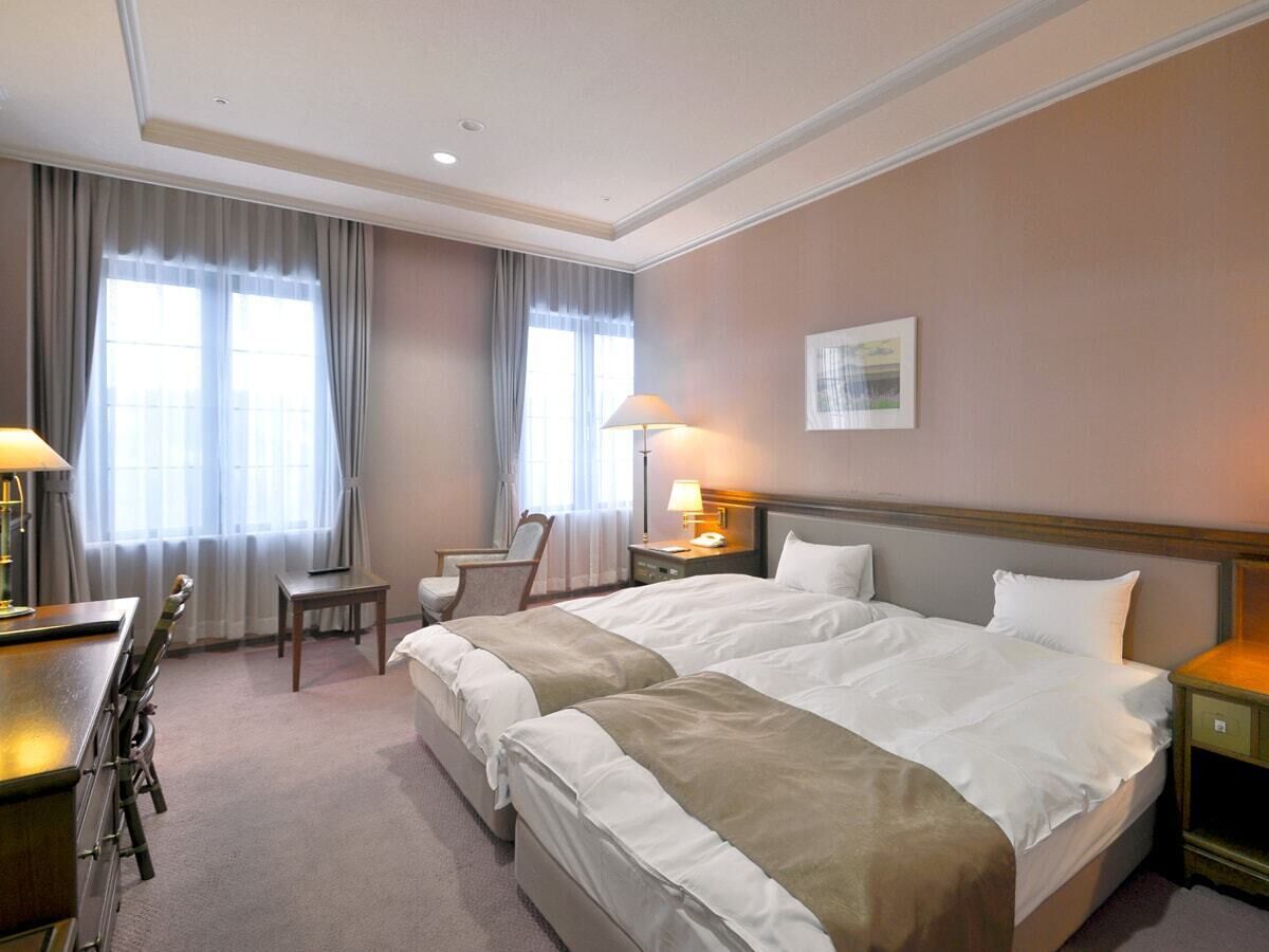 A hotel room with two beds, a desk, a chair, and a lamp.