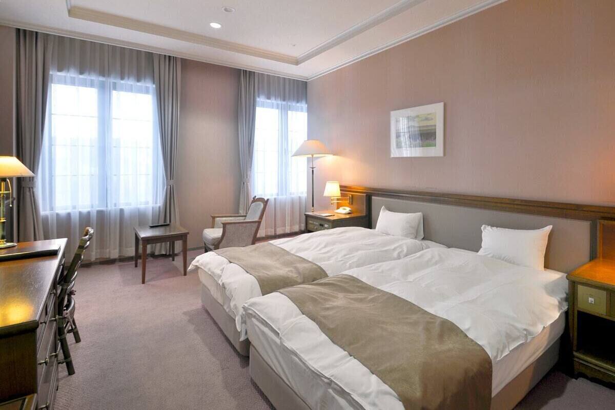 Hotellihuone, jossa on kaksi sänkyä, työpöytä, tuoli ja lamppu.