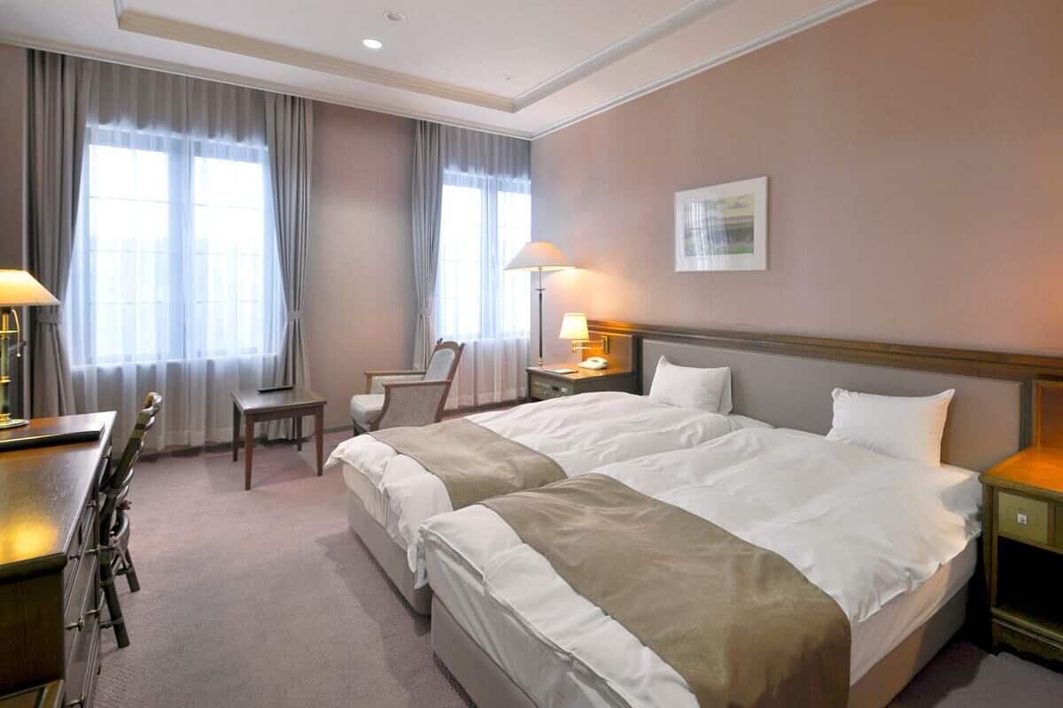 A hotel room with two beds, a desk, a chair, and a lamp.