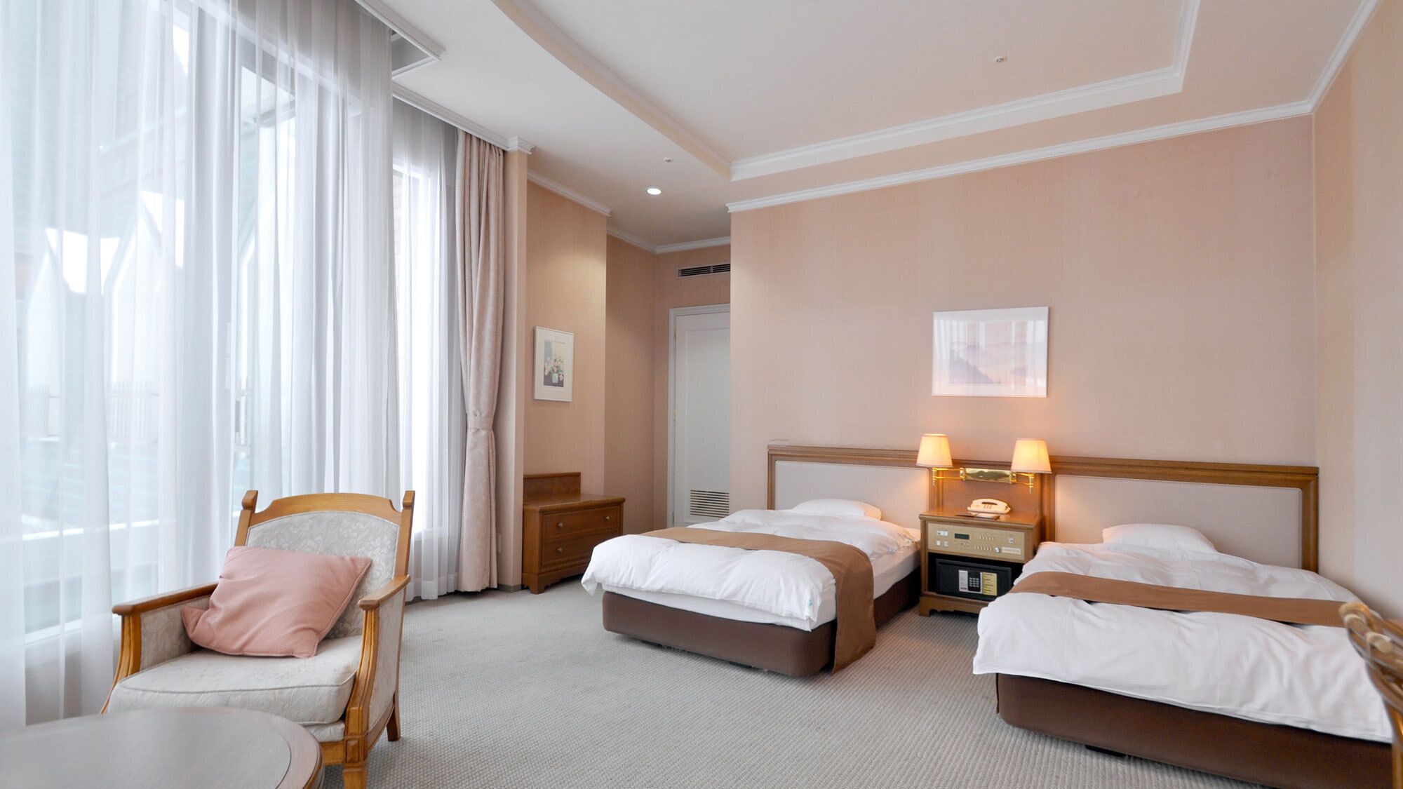 A hotel room with two beds, a chair, a bedside table, a lamp, and a window with curtains.