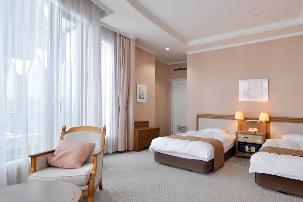 Hotellihuone, jossa on kaksi sänkyä, tuoli, yöpöytä, lamppu ja ikkuna, jossa on verhot.