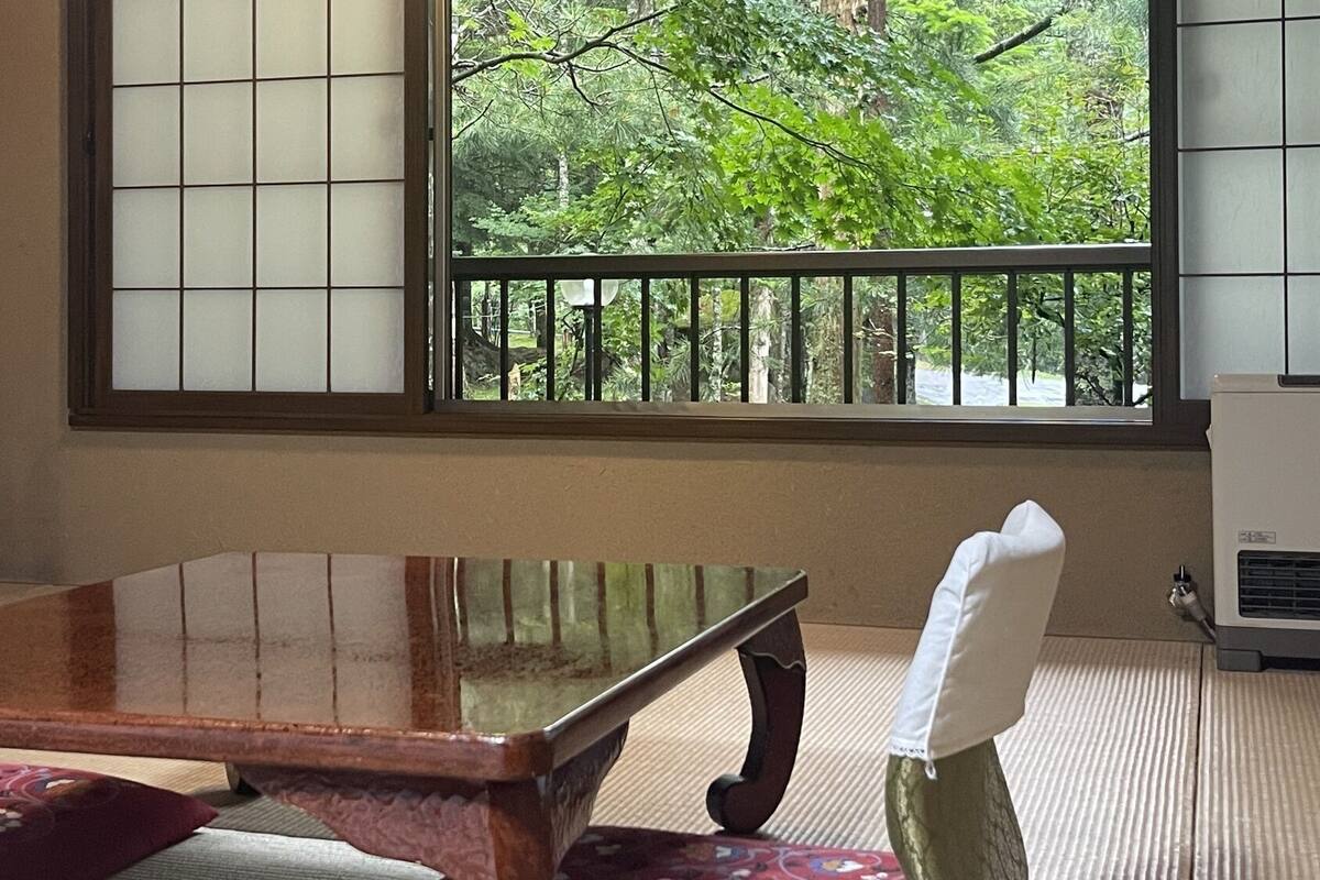 A traditional Japanese room with tatami flooring, a low wooden table, and a view of greenery through a window with a black metal railing.