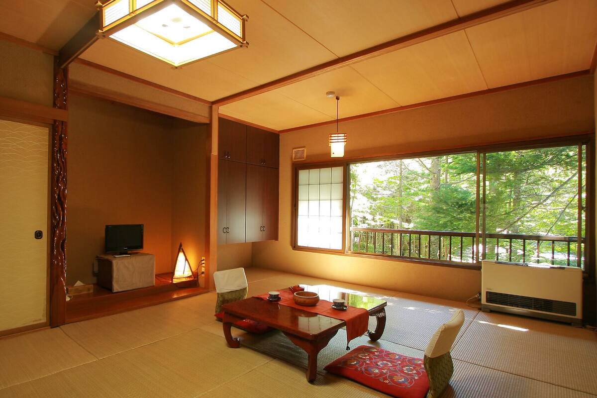 A traditional Japanese room with tatami flooring, a low wooden table, a television on a stand, a sliding door, and a window with a view of trees.