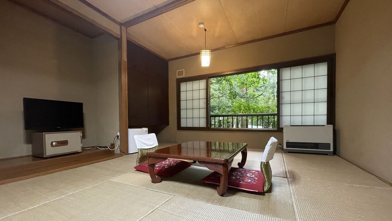 Ein traditioneller japanischer Raum mit Tatami-Böden, einem niedrigen Tisch, einem Fernseher und großen Fenstern mit Blick auf Grünflächen.