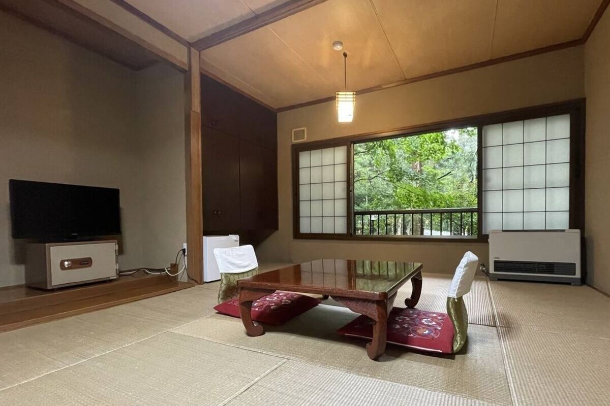 A traditional Japanese room with tatami flooring, a low table, a television, and large windows with a view of greenery.