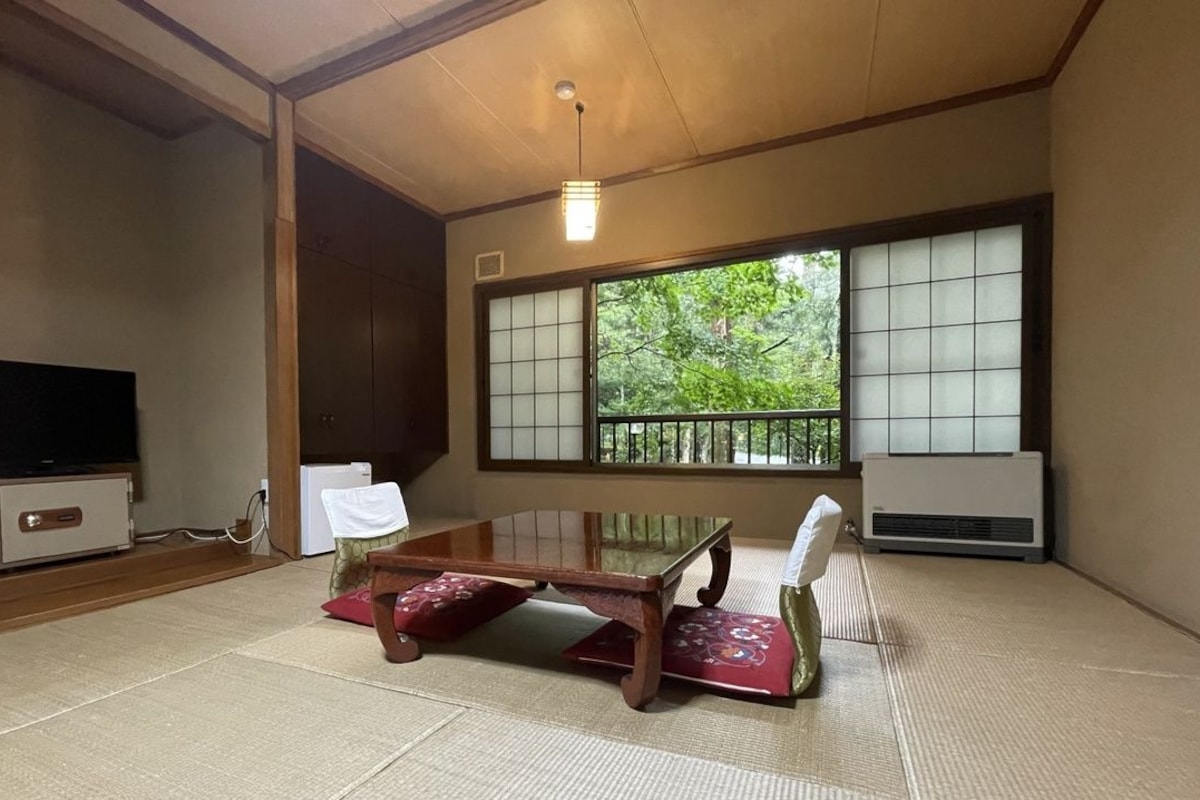 Een traditionele Japanse kamer met tatami vloeren, een lage tafel, een televisie en grote ramen met uitzicht op groen.