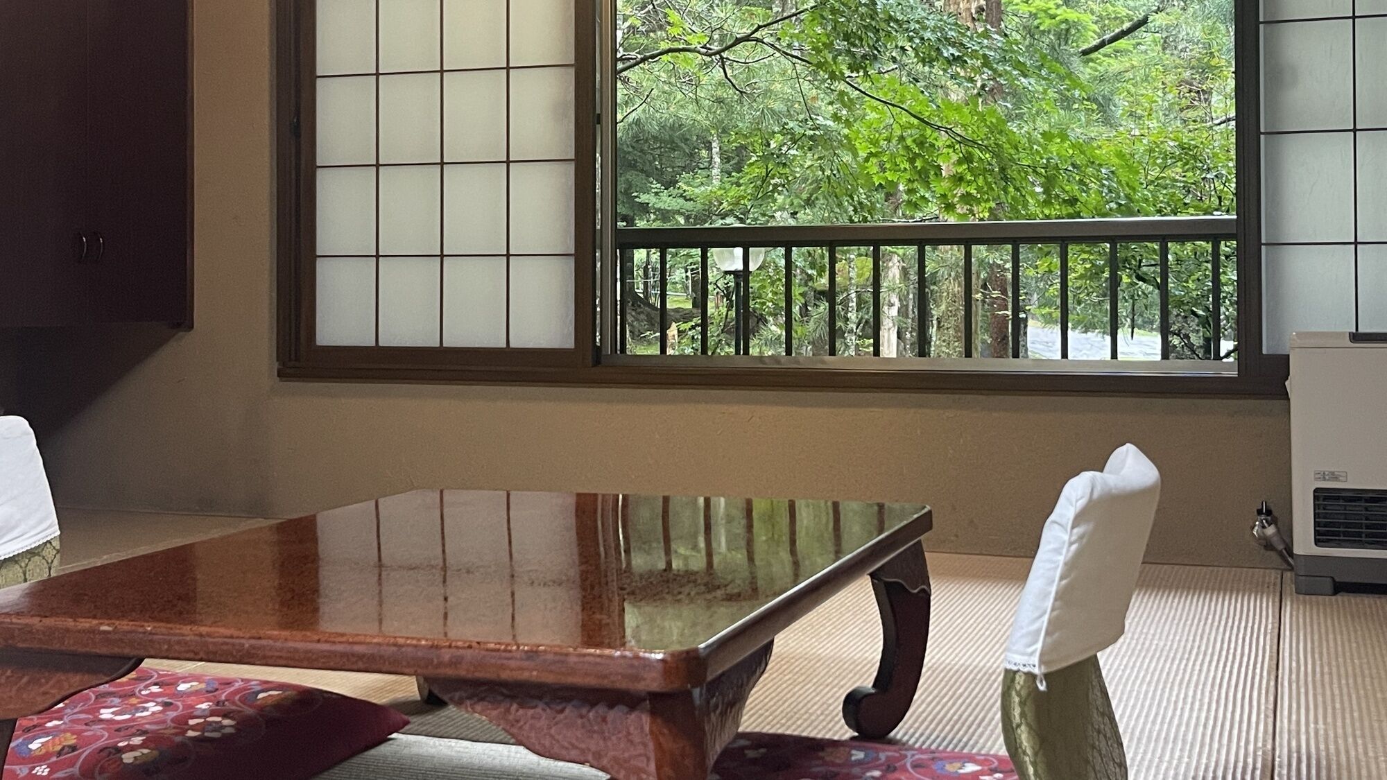 Ein traditioneller japanischer Raum mit Tatami-Bodenbelag, einem niedrigen Holztisch und Blick ins Grüne durch ein Fenster mit schwarzem Metallgeländer.