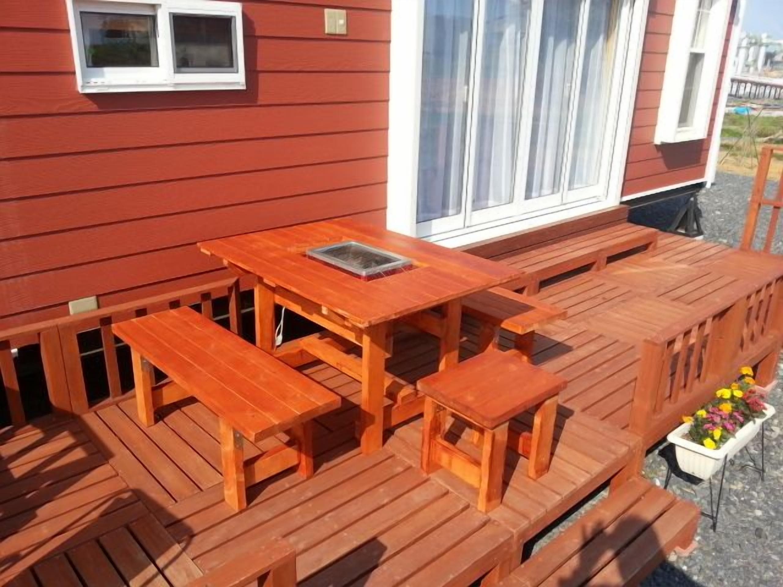 Una terraza de madera con una mesa, bancos, un ventanal y una jardinera con flores.