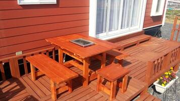 Una terraza de madera con una mesa, bancos, un ventanal y una jardinera con flores.