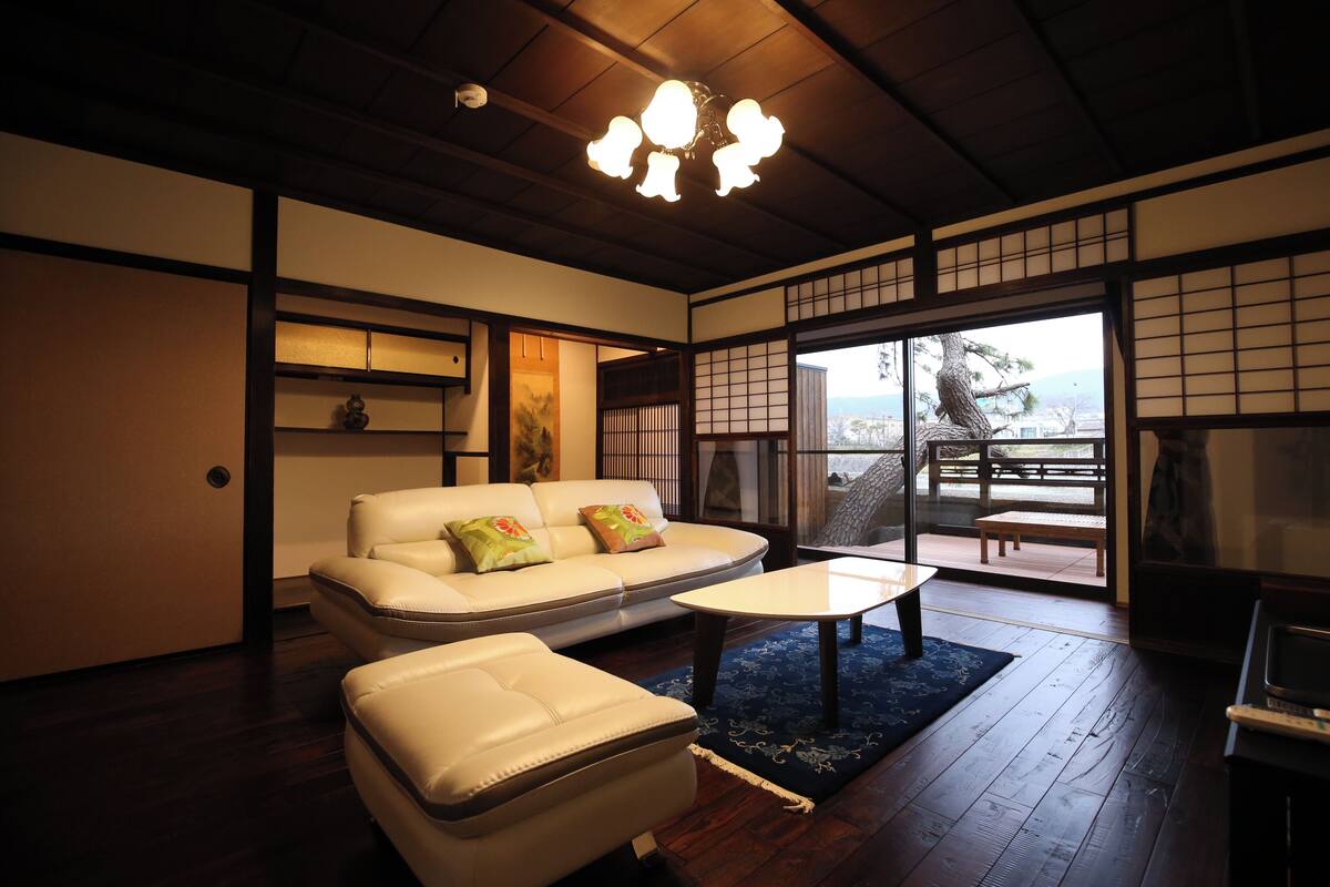 Um quarto tradicional japonês com piso de tatame, teto de madeira baixo, um sofá com almofadas, uma mesa pequena e uma porta de vidro deslizante que dá para uma área externa.