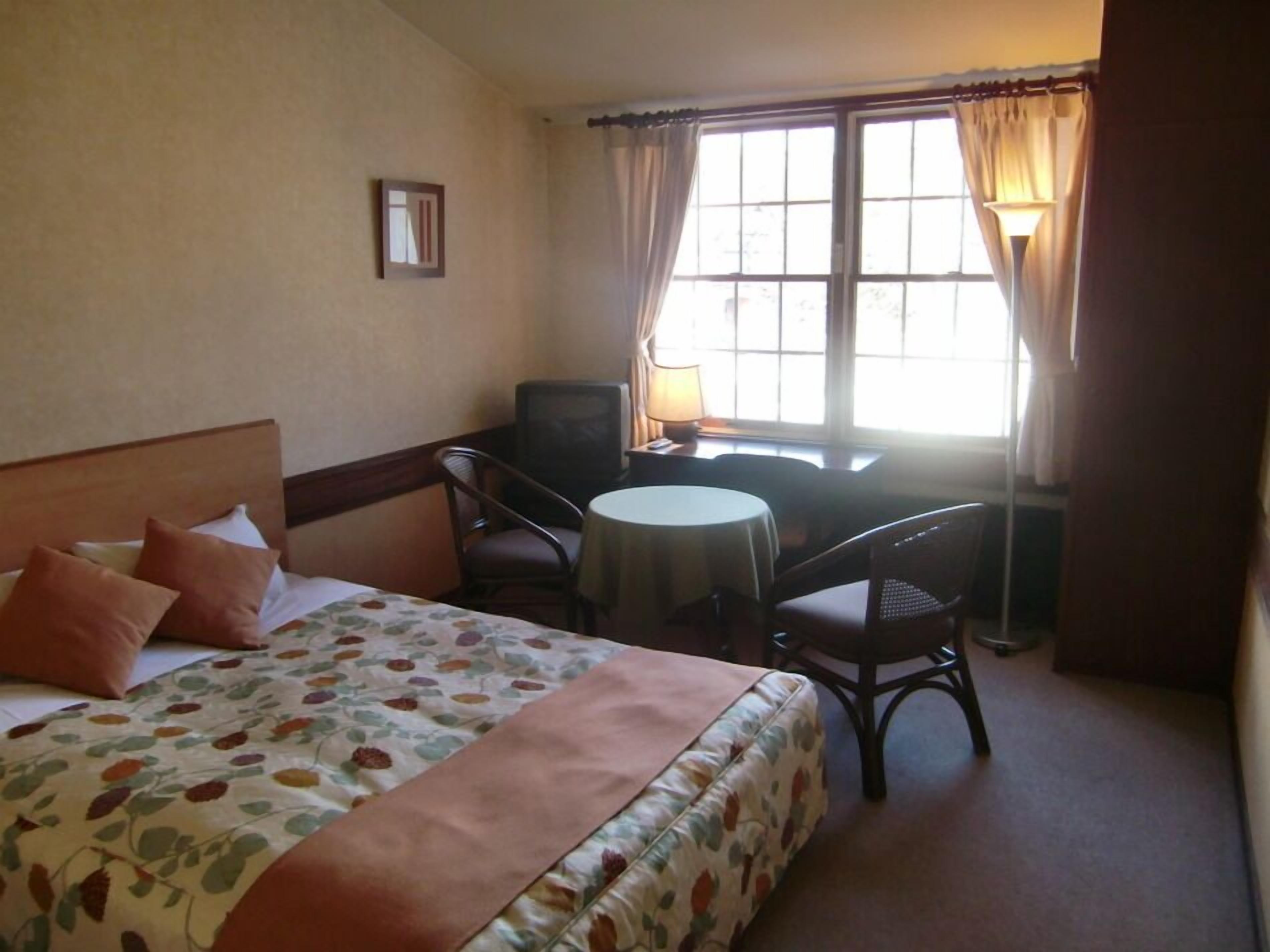 Habitación de hotel con cama, una mesita redonda, dos sillas, televisión y ventana con cortinas.