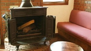 Una estufa de leña con revestimiento de ladrillo, un taburete de madera y un sofá rojo en una habitación con paredes de ladrillo y una ventana.