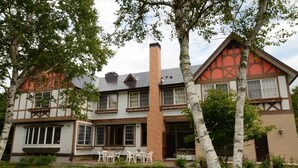 Edificio de dos plantas con chimenea de ladrillo, ventanas amplias y un porche con mobiliario exterior.