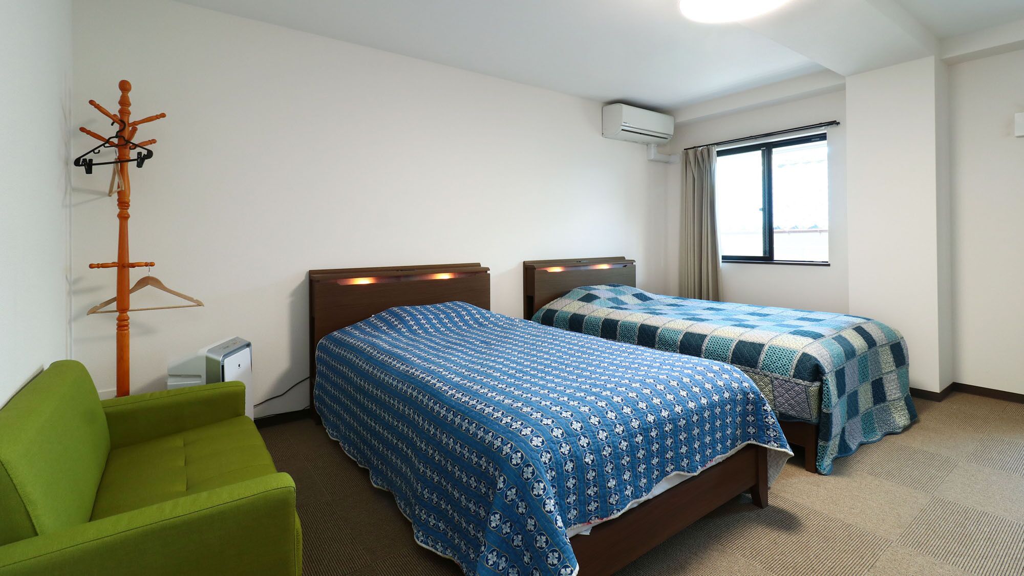 Habitación de hotel con dos camas, una silla verde, un equipo de aire acondicionado y una ventana con cortinas.